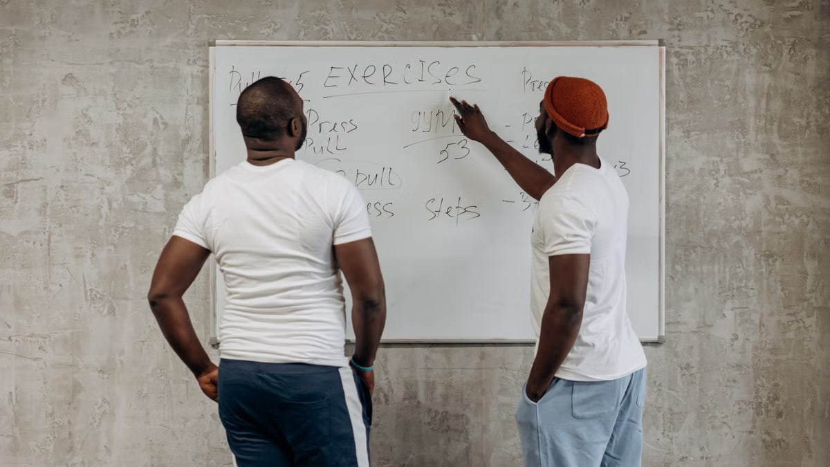 Two adult men planning their gym exercises on a whiteboard in a modern setting, emphasizing fitness and health.